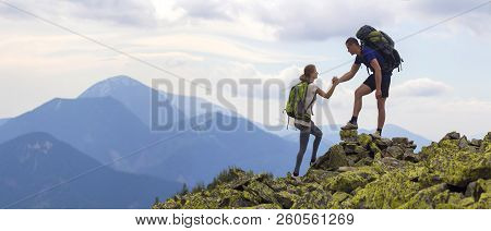 Young Tourists With Backpacks, Athletic Boy Helps Slim Girl To Clime Rocky Mountain Top Against Brig