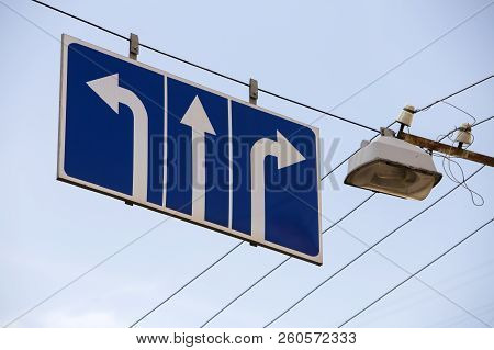 Close-up Of Road Sign Attached To Trolley Line Near The Lamp High Over Street With Three White Arrow
