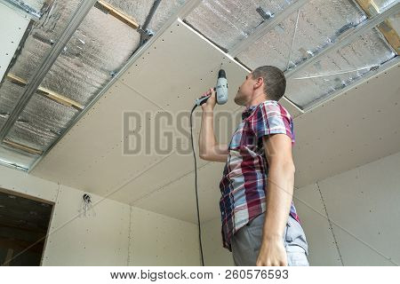 Young Man In Usual Clothing And Work Gloves Fixing Drywall Suspended Ceiling To Metal Frame Using El