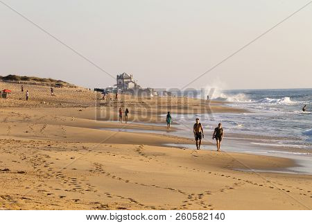 A Man And A Woman Walk In The Sand, Near The Ocean, With The Senhor Da Pedra Chapel In The Backgroun
