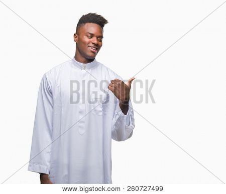 Young arabic african man wearing traditional djellaba over isolated background smiling with happy face looking and pointing to the side with thumb up.