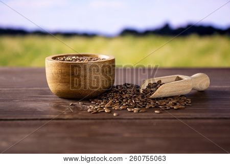 Lot Of Whole Fresh Brown Flaxseeds In A Scoop With Wooden Bowl With Green Wheat Field In Background