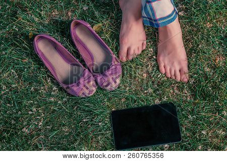 Girl Sitting Crosslegged On Grass With Her Shoes And Tablet Computer Near, Top View