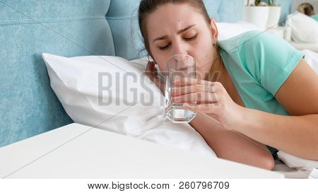 Portrait Of Young Woman Feeling Unwell Lying In Bed And Dirnking Water