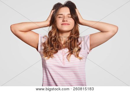 Photo Of Pleased Realxed Young Woman Keeps Both Hands On Head, Eyes Closed, Dressed In Casual Nightw