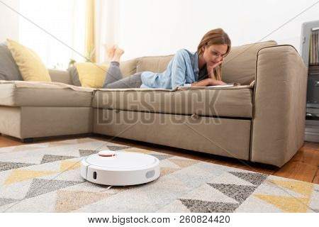 Robotic Vacuum Cleaner Cleaning The Room While Woman Relaxing On Sofa. Woman Controlling Vacuum With