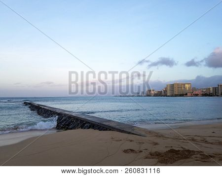 Rock Pier Leading Into Pacific Ocean At Dusk Off The Coast Of Waikiki In Oahu, Hawaii With Hotels An