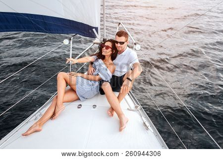 Happy Young Woman Is Lying On Yacht And Point Forward. She Smiles. Guy Sits On Bow Of Yacht With His