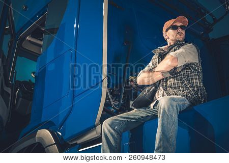 Young Caucasian Trucker Wearing Sunglasses Taking Break On The Truck Stop. Blue Modern Semi Truck.