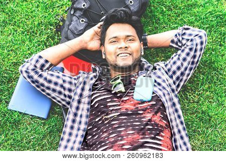 Young Indian Man Daydreaming At Park Listening Music By Phone With Dreamy Facial Expression - Happy 