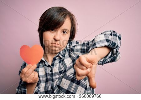 Young down syndrome romantic woman holding red heart paper shape over pink background with angry face, negative sign showing dislike with thumbs down, rejection concept