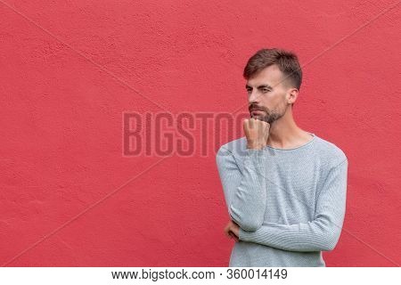 Man In Thoughtful Pose With Hand On Red Wall Background With Copy Space. Standing Thinking Male. Min