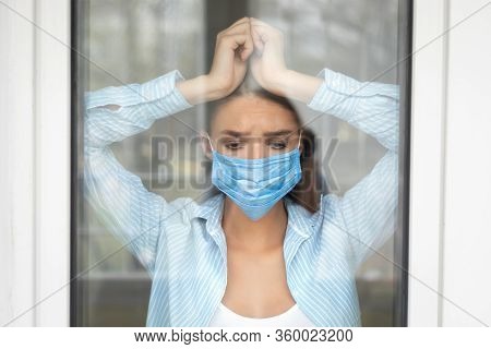 Coronavirus Quarantine. Depressed Woman Standing Near Windor At Home Looking Outside, Wearing Protec