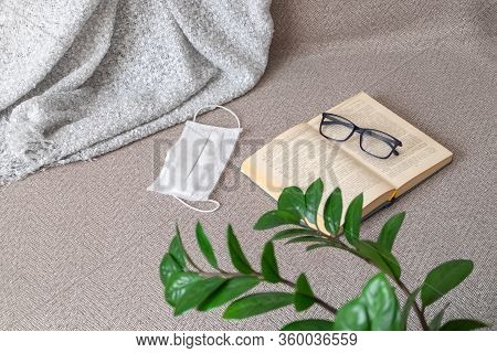 An Open Book, Glasses, A Medical Mask And Plaid Lie On The Sofa Near A Green Houseplant. Covid-19, S