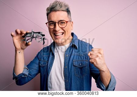 Young modern optical optomestrist man holding optometrical glasses over pink background screaming proud and celebrating victory and success very excited, cheering emotion