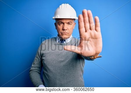 Middle age handsome grey-haired engineer man wearing safety helmet over blue background doing stop sing with palm of the hand. Warning expression with negative and serious gesture on the face.