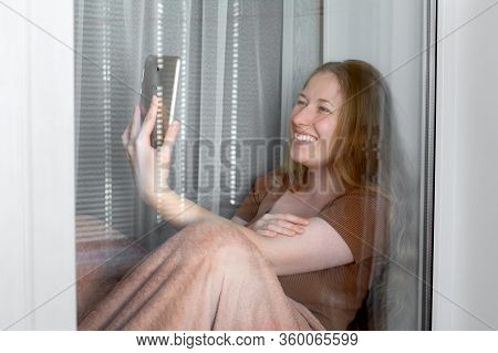A Happy Smile Girl Sits On A Windowsill By The Window, Wrapped In A Blanket And Holds A Tablet In Ha