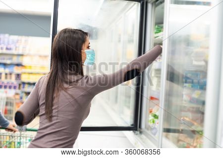 Masked woman taking deep frozen food from a freezer in a spuermarket during coronavirus pandemic