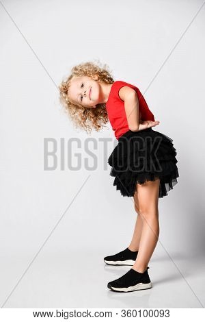 Playful Curly Hair Blonde Kid Girl In Red T-shirt, Black Skirt And Sneakers Poses Side To Us Bending