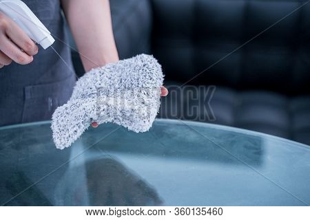 Young Woman Maid In Apron Is Cleaning, Wiping Down Office Glass Table Surface With Spray Bottle Clea