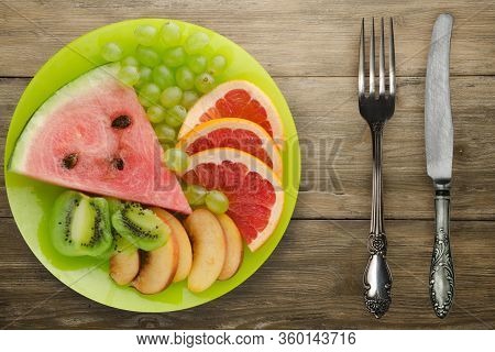 Sliced Fruit On A Borwn Wooden Background. Sliced Fruit On A Light Green Plate Fork And Knife Top Vi