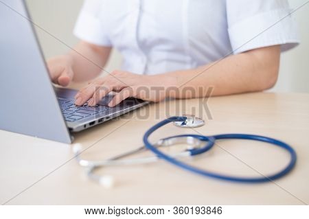 Unrecognizable Female Doctor In Uniform Uses A Laptop. Faceless Medical Worker Writes A Report On Th