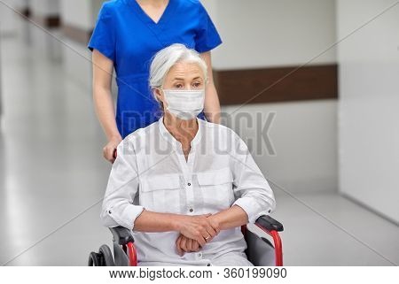 medicine, health safety and pandemic concept - nurse carrying senior woman patient in wheelchair wearing face protective medical mask for protection from virus disease at hospital corridor
