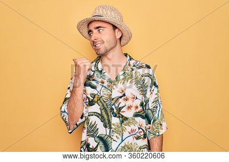 Young handsome man with blue eyes on vacation wearing summer florar shirt and hat smiling with happy face looking and pointing to the side with thumb up.