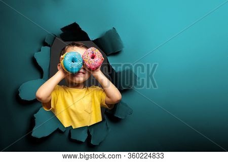 Happy cute boy is having fun played with donuts on black background wall. Bright photo of a boy. Colored donuts