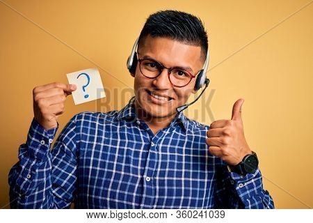 Young latin call center agent man holding question mark reminder paper using headset happy with big smile doing ok sign, thumb up with fingers, excellent sign