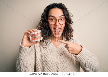 Young beautiful woman with curly hair holding plastic teeth with dental baces very happy pointing with hand and finger