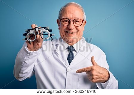 Senior grey haired optic doctor man holding optometrist eyeglasses over blue background with surprise face pointing finger to himself