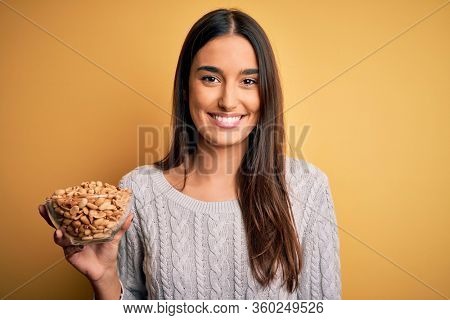 Young beautiful brunette woman holding bowl with peanuts over isolated yellow background with a happy face standing and smiling with a confident smile showing teeth