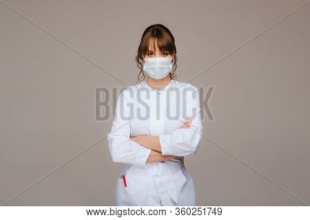Beautiful Young Girl In A White Medical Coat, Medical Mask And Arms Crossed On Her Chest,