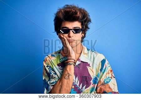 Young handsome man wearing thug life glasses standing over isolated blue background thinking looking tired and bored with depression problems with crossed arms.