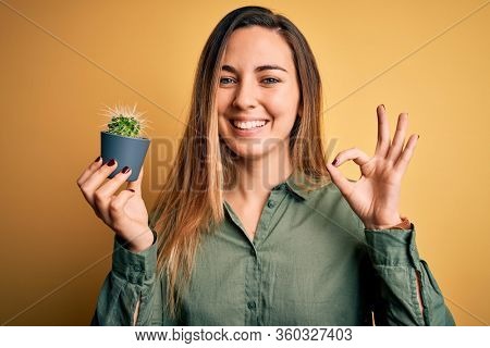 Young beautiful blonde woman with blue eyes holding small cactus pot over yellow bckground doing ok sign with fingers, excellent symbol