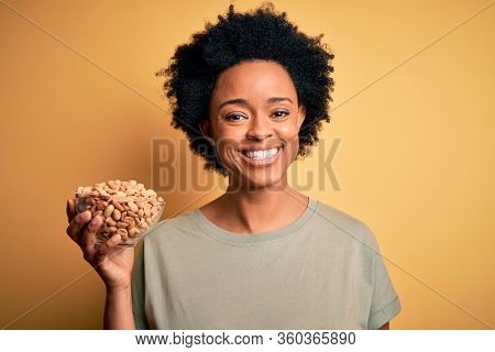 Young African American afro woman with curly hair holding bowl with salty peanuts with a happy face standing and smiling with a confident smile showing teeth
