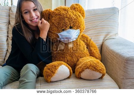 Beautiful Little Girl In A Black Jacket Playing With Her Brown Teddy Bear Who Has Put A Mask To Prot