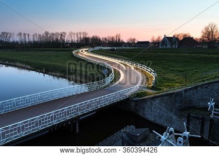 Curved Asphalt Country Road In Dutch Countryside With White Fence Leading Towards An Old Farmhouse I