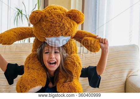 Beautiful Little Girl In A Black Jacket Playing With Her Brown Teddy Bear Who Has Put A Mask To Prot