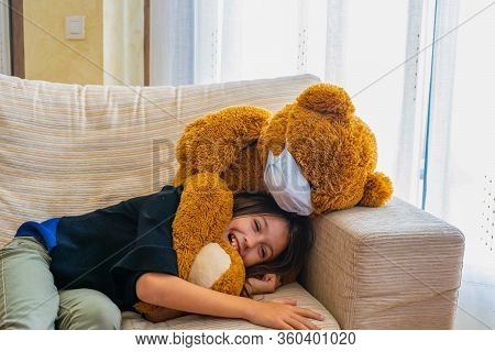 Beautiful Little Girl In A Black Jacket Playing With Her Brown Teddy Bear Who Has Put A Mask To Prot