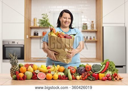 Corpulent woman holding a paper bag with groceries in a kitchen with fruits and vegetables on the table