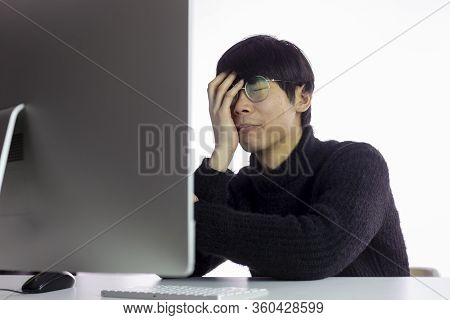 Young Asian Overworked Men In Glasses Sitting In Office, Looks Sick, Tired Of Work. Hand Covers Face