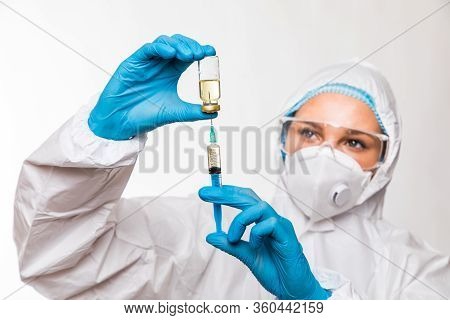 Female Doctor In A Protective White Suit With A Syringe In His Hands And Blue Rubber Gloves Stands I