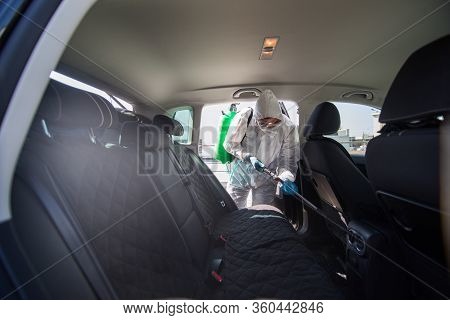 Coronavirus Pandemic. A Disinfector In A Protective Suit And Mask Sprays Disinfectants On Car. Prote