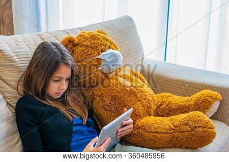 Beautiful Little Girl In A Black Jacket Playing With Her Brown Teddy Bear Who Has Put A Mask To Prot