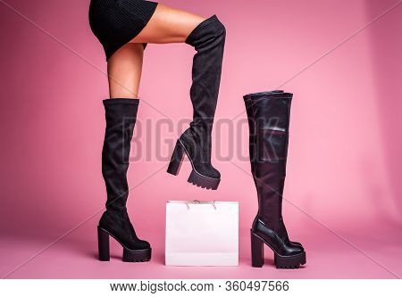 The Girl Stands With Her Foot Lifted Up In Black Jackboots Boots On Her Feet On A Pink Background. I