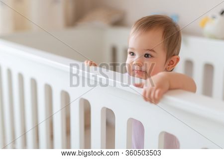 Portrait Of Cute Baby Standing In Crib. Baby Girl Stand Alone In Crib. Happy Baby Girl In White Baby