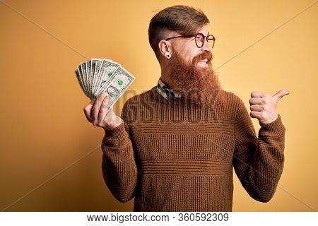 Redhead Irish man with beard holding a bunch of 1 dolla banknotes over yellow background pointing and showing with thumb up to the side with happy face smiling