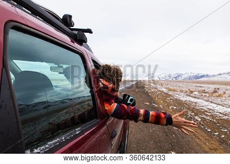 Backview Of Girl In Bright Burgundy Colored Jacket Holding Hand Out Of The Car At Empty Road With Mo